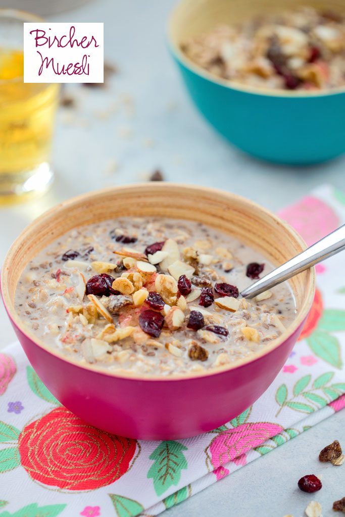 Head-on view of a pink bowl of bircher muesli on a flowered towel with teal bowl, glass of apple juice, and dried berries in the background and recipe title at top