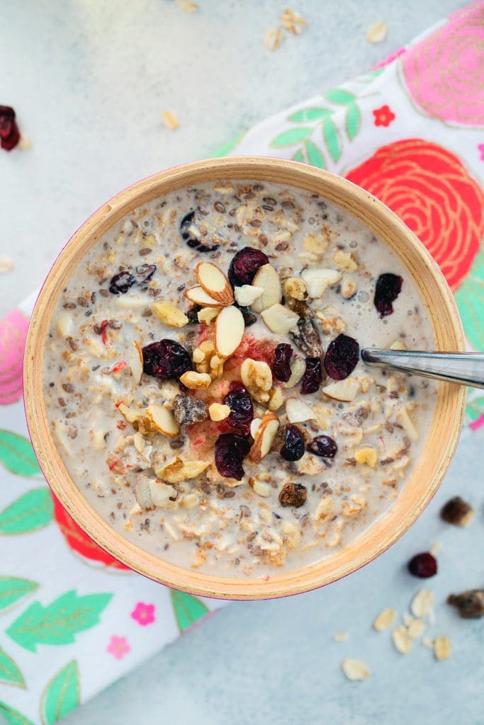 Overhead view of a bowl of bircher muesli topped with dried fruit and nuts on a flowered towel with dried fruits and oats scattered around