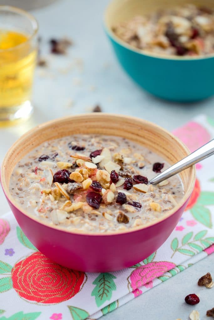 Head-on view of a pink bowl of bircher muesli on a flowered towel with teal bowl, glass of apple juice, and dried berries in the background