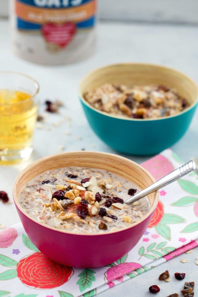 View from a distance of two bowls of bircher muesli topped with dried fruit with glass of apple juice, carton of oats, and dried fruit in the background