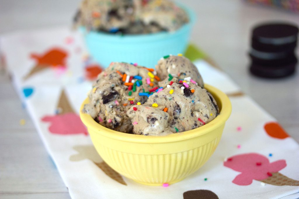 Landscape head-on view of a yellow bowl of coffee birthday cake Oreo ice cream with rainbow sprinkles with second  bowl of ice cream in the background and stack of birthday cake Oreo cookies