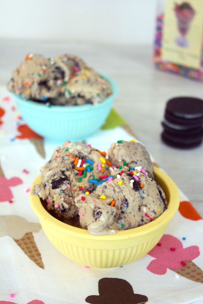 Head-on view of a yellow bowl of coffee birthday cake ice cream with birthday cake Oreos and rainbow sprinkles on a ice cream-themed tea towel with a second bowl of ice cream, Oreo cookie stack, and sprinkles in the background