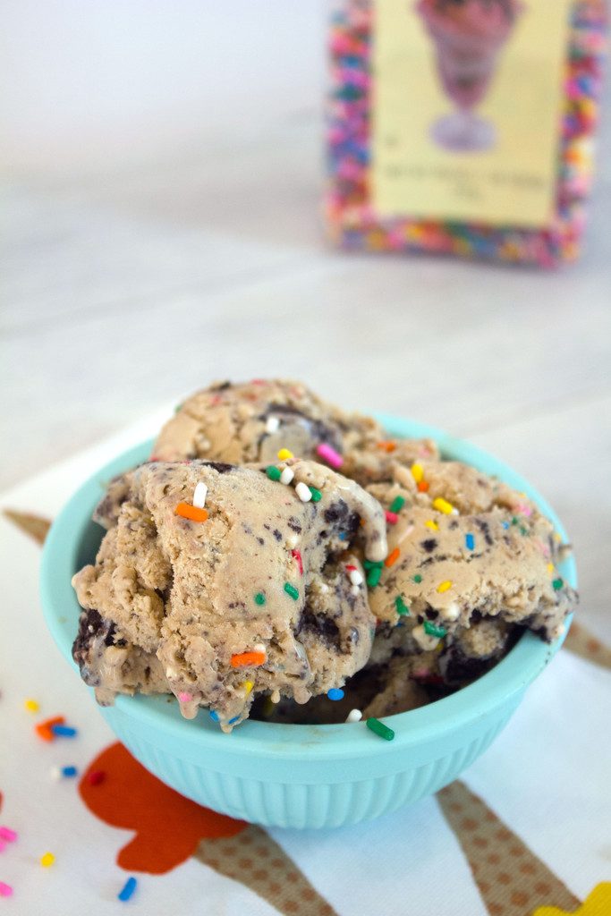 Overhead view of a blue bowl of coffee birthday cake Oreo ice cream with rainbow sprinkles and a large container of sprinkles in the background