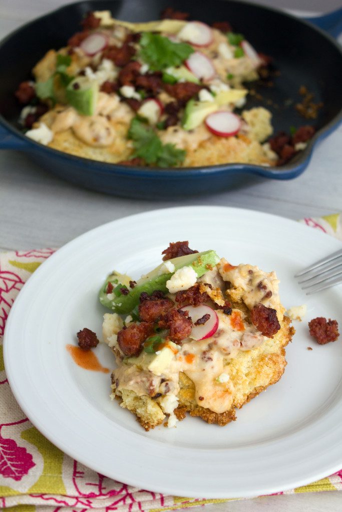 Overhead view of cornmeal biscuit with chorizo gravy topped with chorizo, avocado, radishes, and cheese on a plate with skillet in background