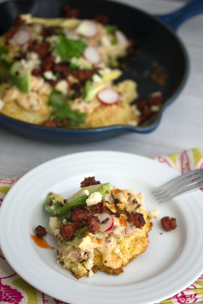 Plate with a cornmeal biscuit with chorizo gravy with avocado, cheese, and radishes with skillet of biscuits and gravy in background