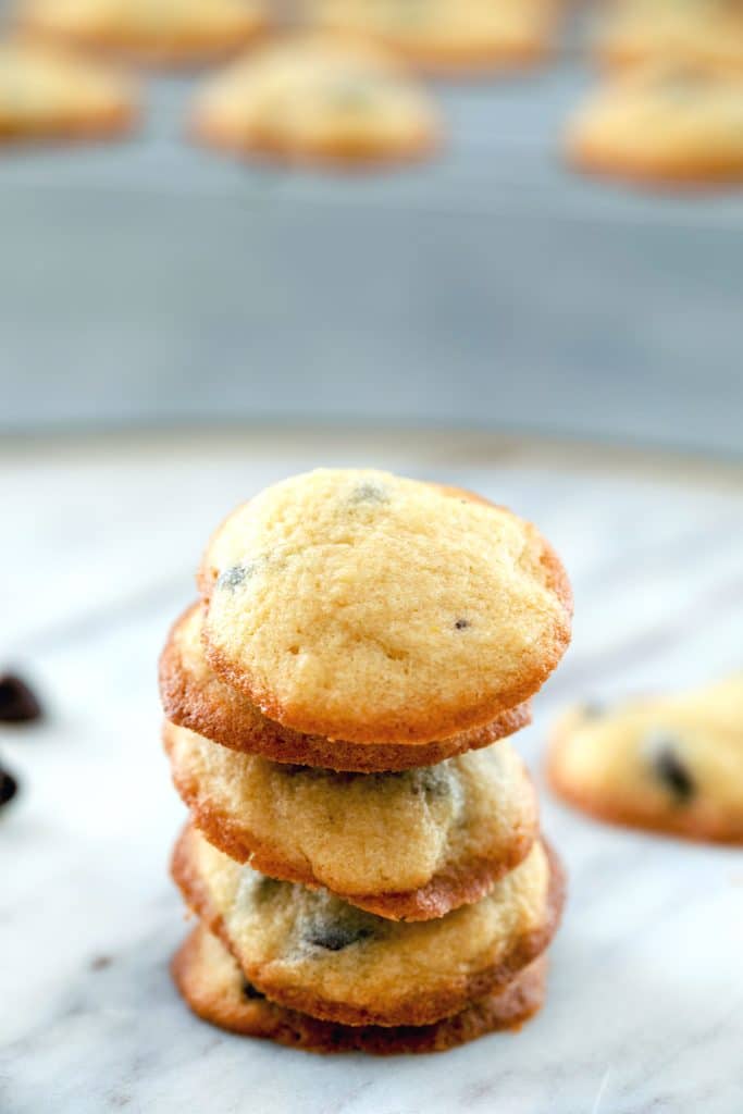 Overhead view of bite-size chocolate chip cookies stacked up on marble surface with baking rack with more cookies in the background