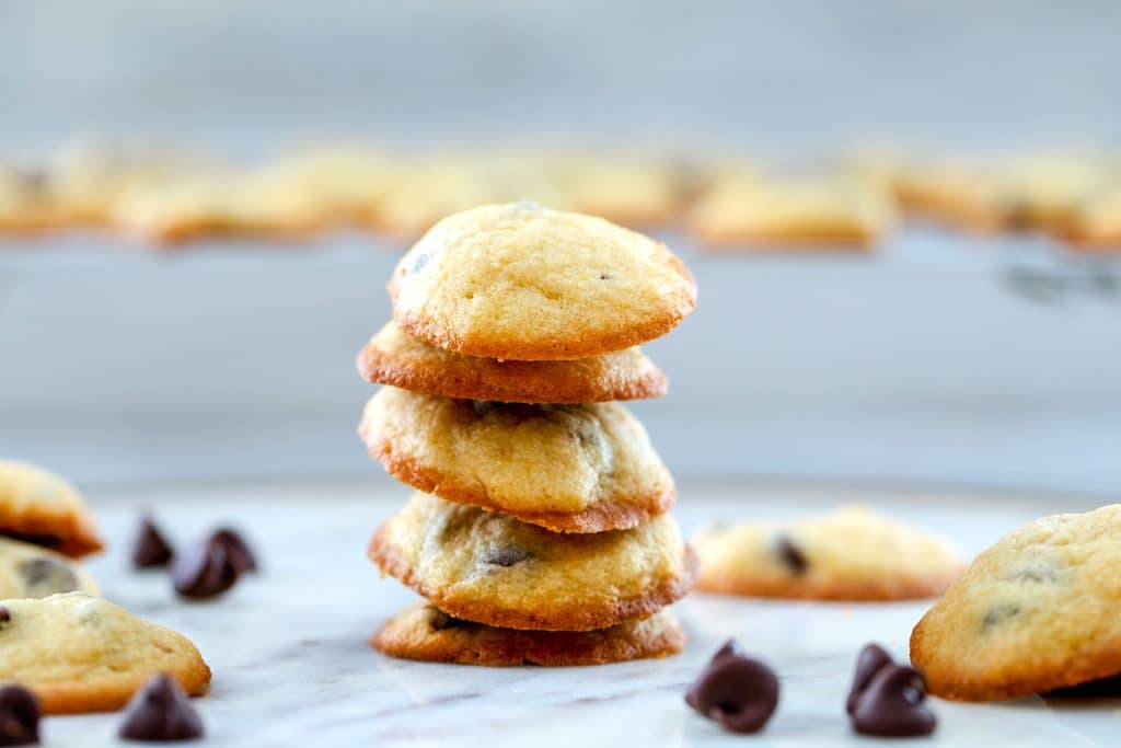 Landscape head-on view of five bite-size chocolate chip cookies stacked up on marble surface surrounded by chocolate chips and more cookies and baking rack with more cookies in background