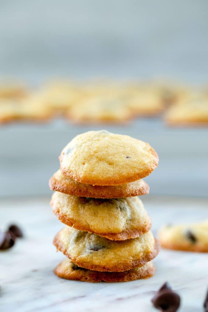 Head-on view of five bite-size chocolate chip cookies stacked on top of each other with chocolate chips and more cookies in the background