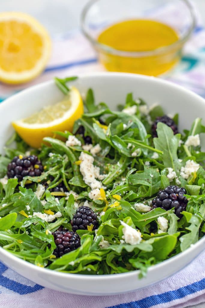 Head-on view of blackberry feta salad with arugula in a white bowl with lemon wedge and dressing and lemon in the background