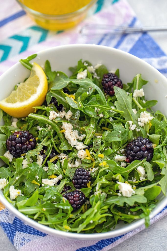 Overhead view of blackberry feta salad with arugula and lemon wedge in a white bowl on a lavender napkin with yellow dressing and small whisk in the background