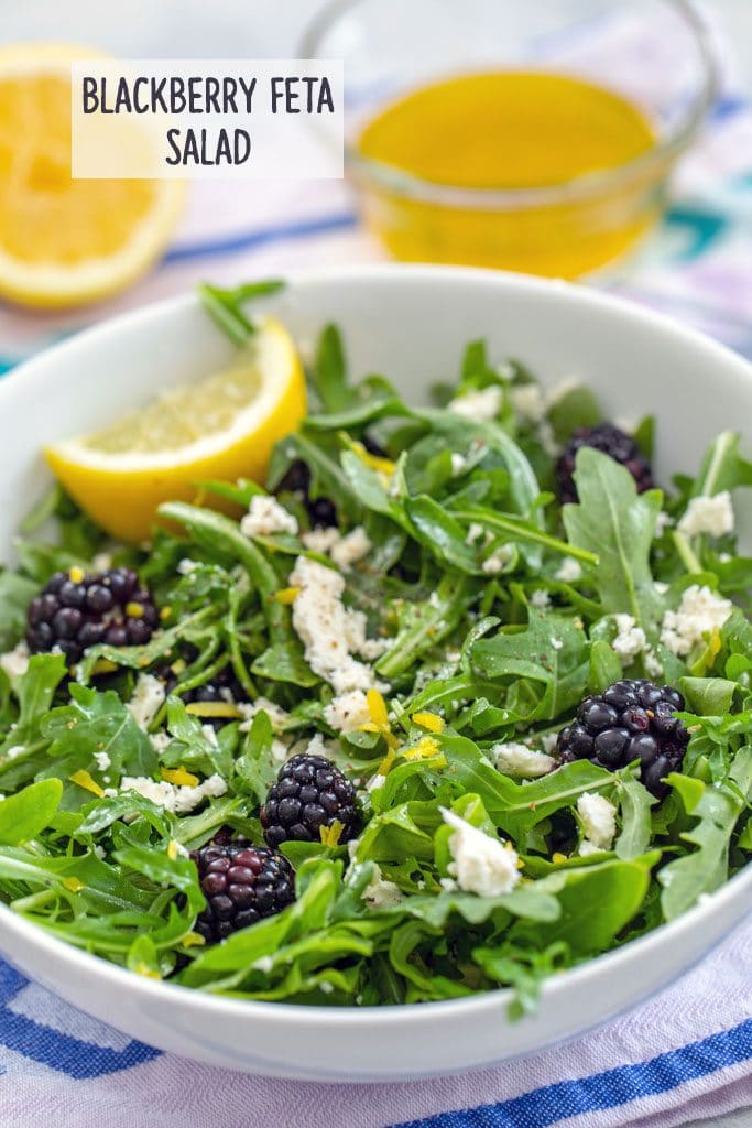 Head-on view of blackberry feta salad with arugula in a white bowl with lemon wedge and dressing and lemon in the background and "Blackberry Feta Salad" text on top