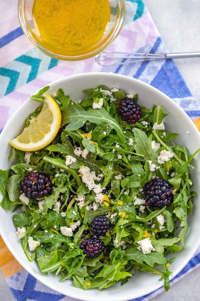 Diret overhead view of blackberry feta salad with a lime wedge in a white bowl on a colorful napkin with a bowl of lemon salad dressing and a small whisk in the background