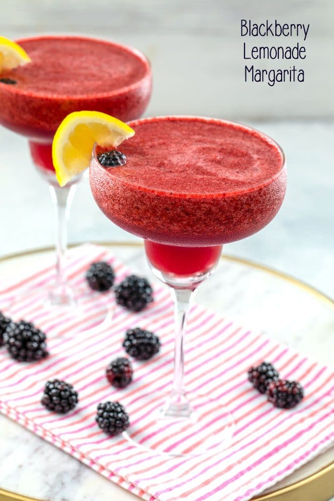 Head-on view of two blackberry lemonade margaritas with lemon garnish on a white marble tray with pink striped towel and blackberries scattered around and "Blackberry Lemonade Margarita" text at top