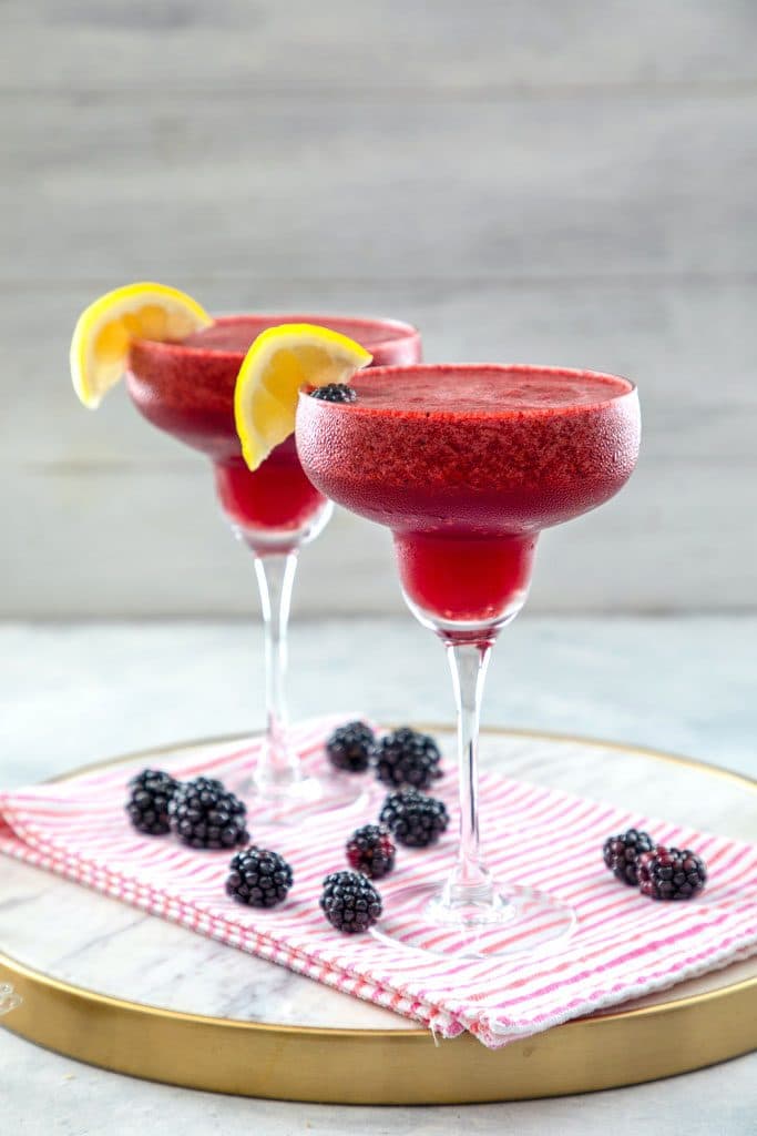 Straight-on view from a distance of blackberry lemonade margaritas with lemon garnishes on a marble tray with pink striped towel and blackberries all around