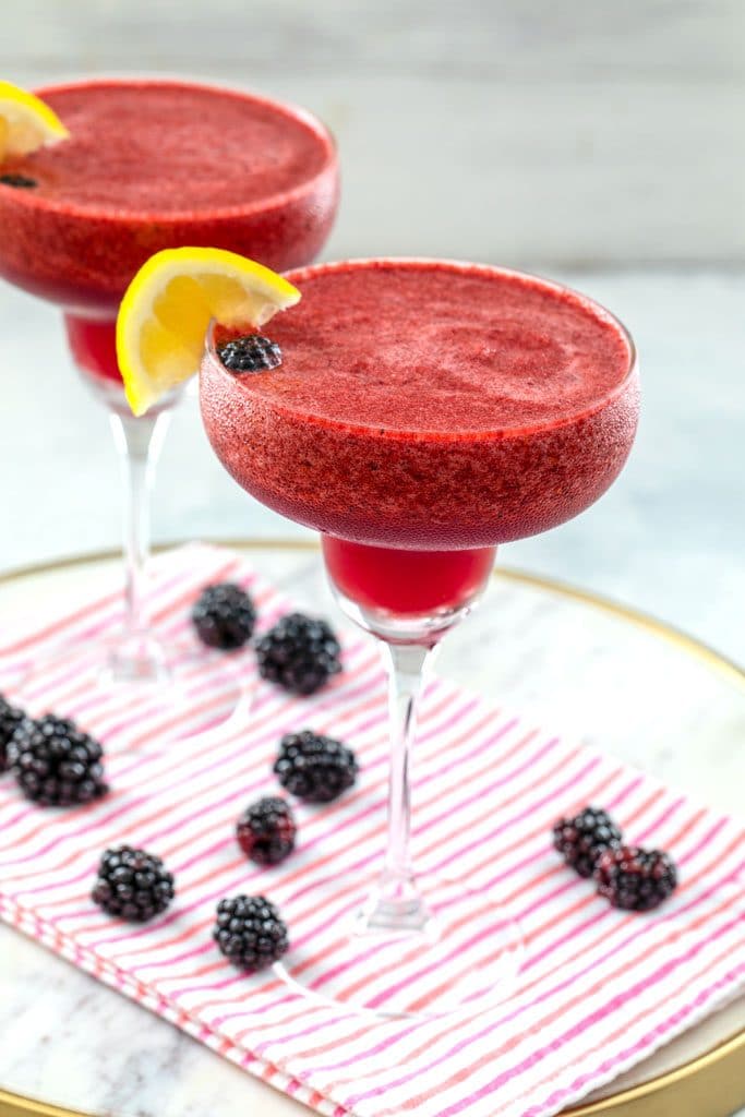 Head-on view of two blackberry lemonade margaritas with lemon garnish on a white marble tray with pink striped towel and blackberries scattered around