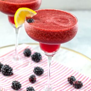 Head-on view of two blackberry lemonade margaritas with lemon garnish on a white marble tray with pink striped towel and blackberries scattered around and "Blackberry Lemonade Margarita" text at top