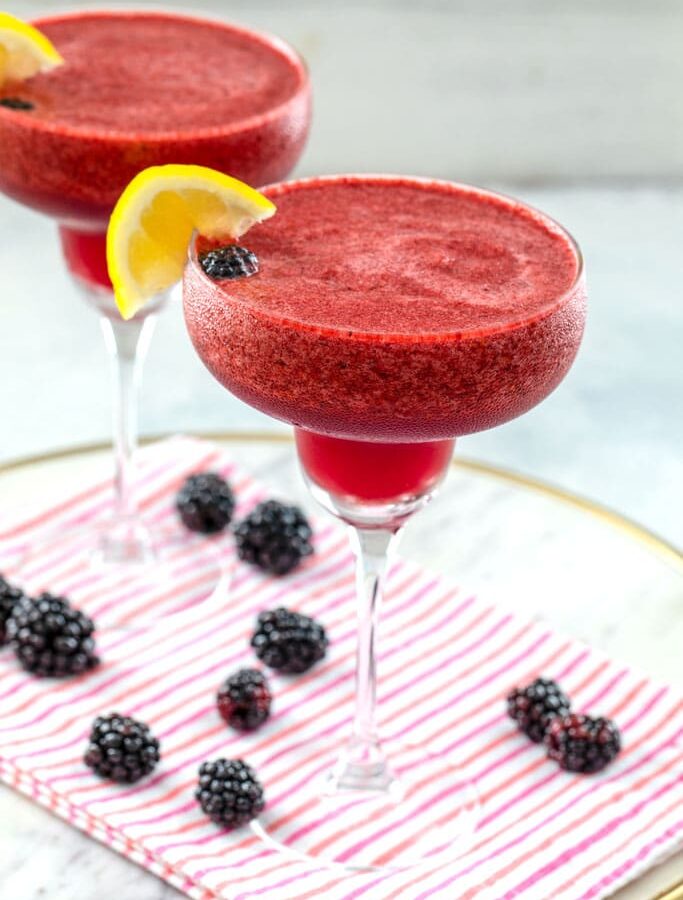 Head-on view of two blackberry lemonade margaritas with lemon garnish on a white marble tray with pink striped towel and blackberries scattered around and "Blackberry Lemonade Margarita" text at top
