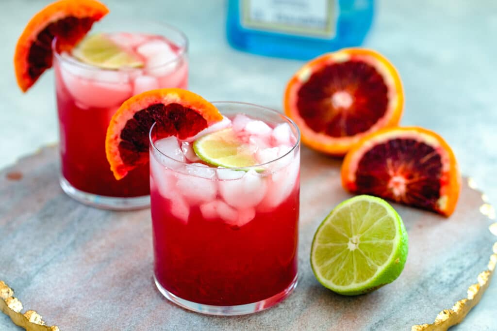 Landscape head-on view or two blood orange gin cocktails on a marbled tray with lime and blood orange halves with bottle of gin in the background