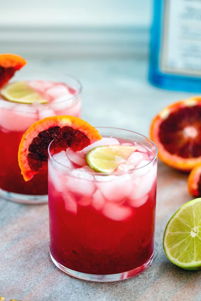 Head-on closeup view of a blood orange lime rickey cocktail with second cocktail, blood orange and lime halves, and bottle of gin in background