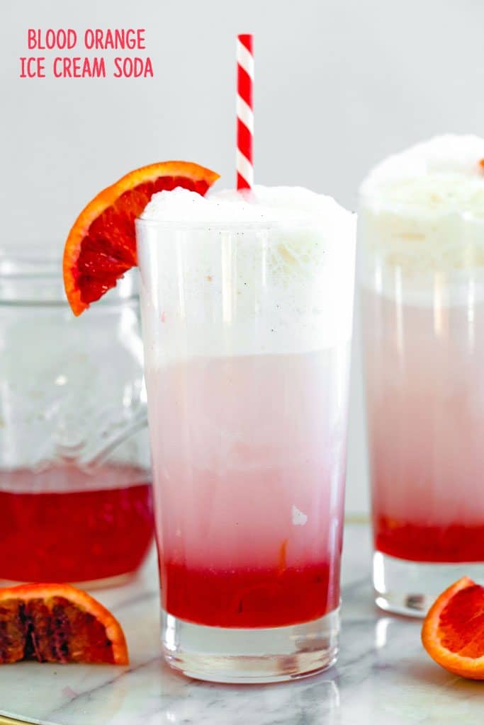 Head-on view of blood orange ice cream soda in a tall glass with red and white straw and blood orange wedge garnish with jar of simple syrup and second ice cream soda in the background and recipe title at top