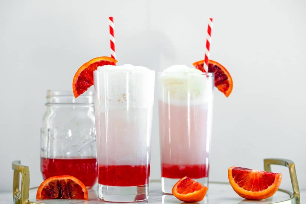 Landscape head-on view of two blood orange ice cream sodas in tall glasses on a marble tray with blood orange wedges and jar of blood orange simple syrup in background