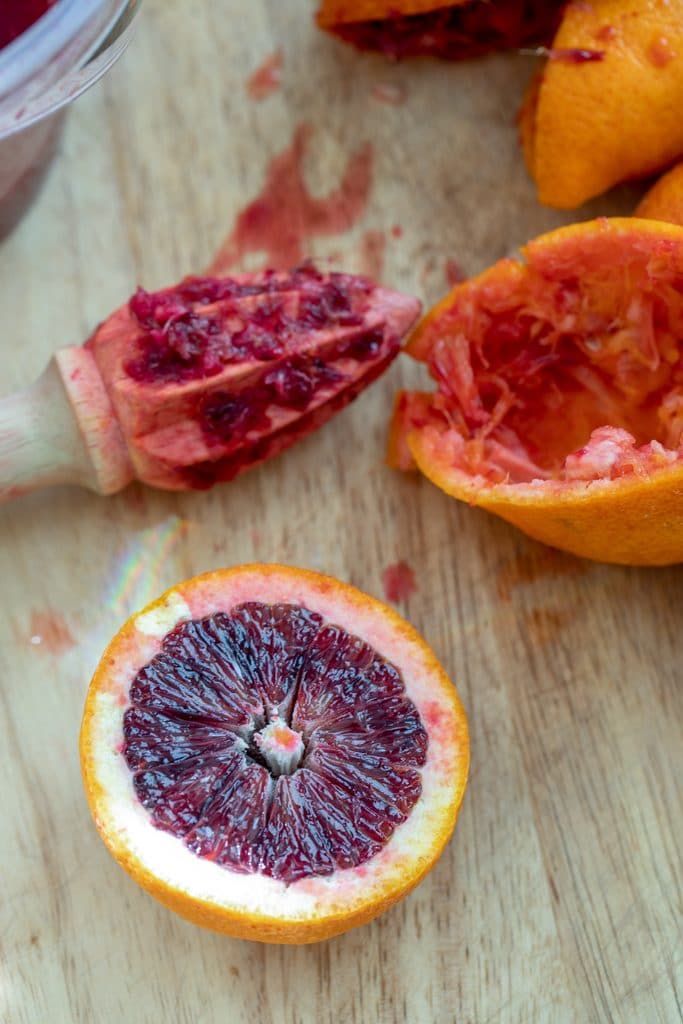 A blood orange half on a cutting board with citrus reamer and juice blood orange halves in the background