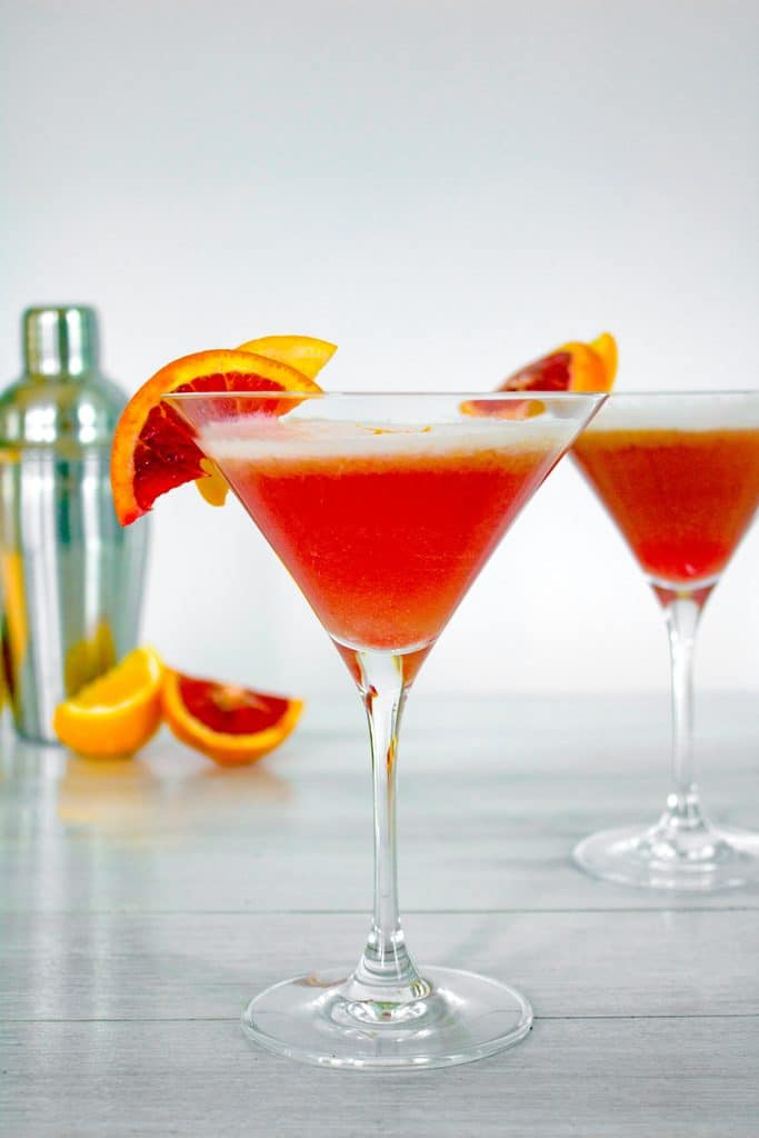 Head-on view of blood orange meyer lemon gin sour in a martini glass with blood orange slice garnish with second cocktail, cocktail shaker, and blood orange slices in the background 