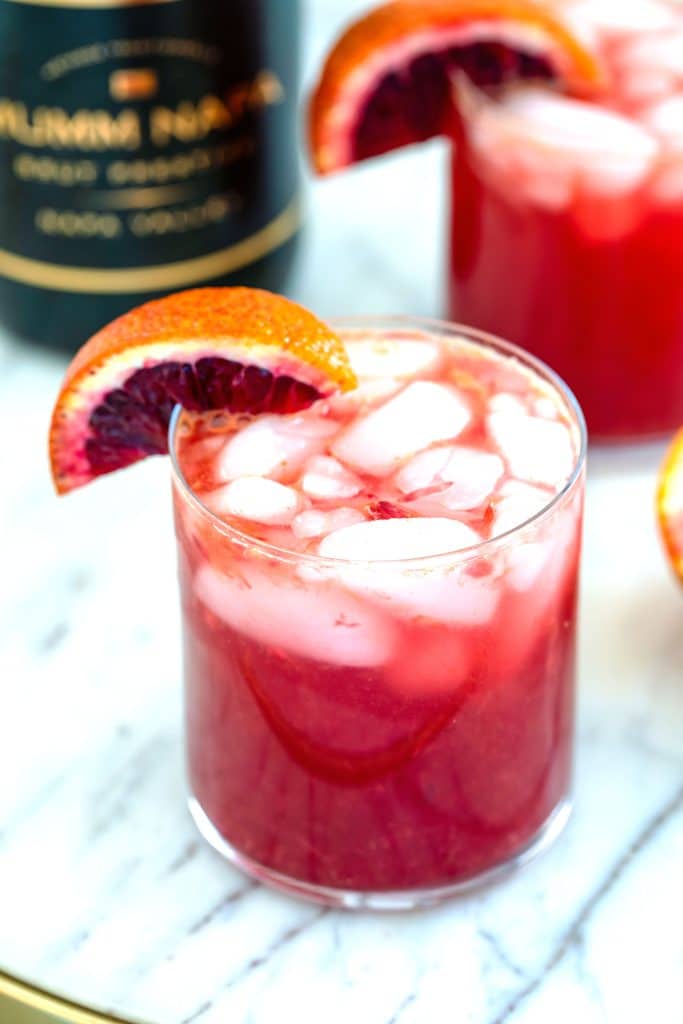 Overhead view of a blood orange cocktail on a marble surface with ice and a blood orange wedge garnish with bottle of champagne and second cocktail in background