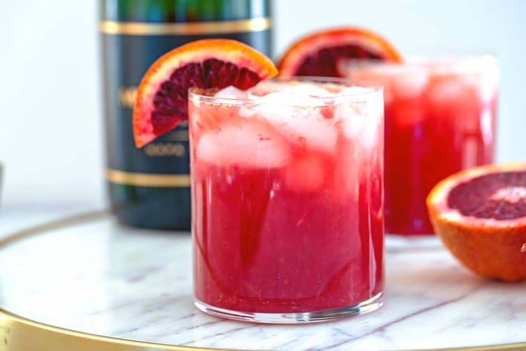 Landscape head-on closeup of a blood orange cocktail on a marble tray with bottle of champagne, second cocktail, and blood orange half in the background