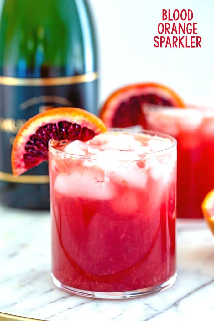 Head-on view of a blood orange cocktail with blood orange wedge garnish on a marble surface with a second cocktail and bottle of champagne in the background and recipe title at top