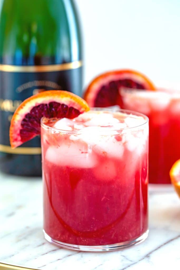 Head-on view of a blood orange cocktail with blood orange wedge garnish on a marble surface with a second cocktail and bottle of champagne in the background