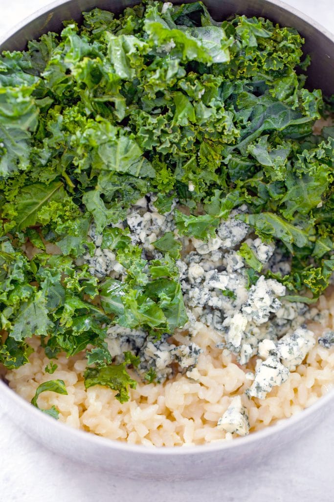 Overhead view of saucepan with arborio rice topped with crumbled blue cheese, and chopped kale leaves 
