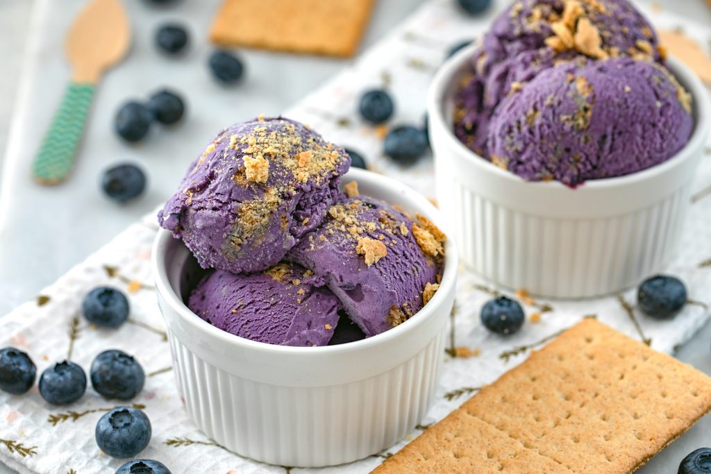 Landscape view of two white bowl of blueberry cheesecake ice cream topped with crushed graham crackers with blueberries, graham crackers, and a wooden spoon