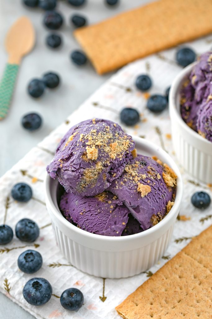 Head-on view of a white bowl of blueberry cheesecake ice cream stopped with crushed graham crackers and surrounded by blueberries and graham crackers 