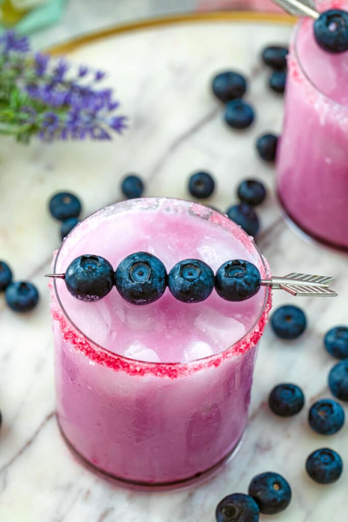 Overhead view of a blueberry coconut margarita with fresh blueberry garnish and blueberries scattered all around