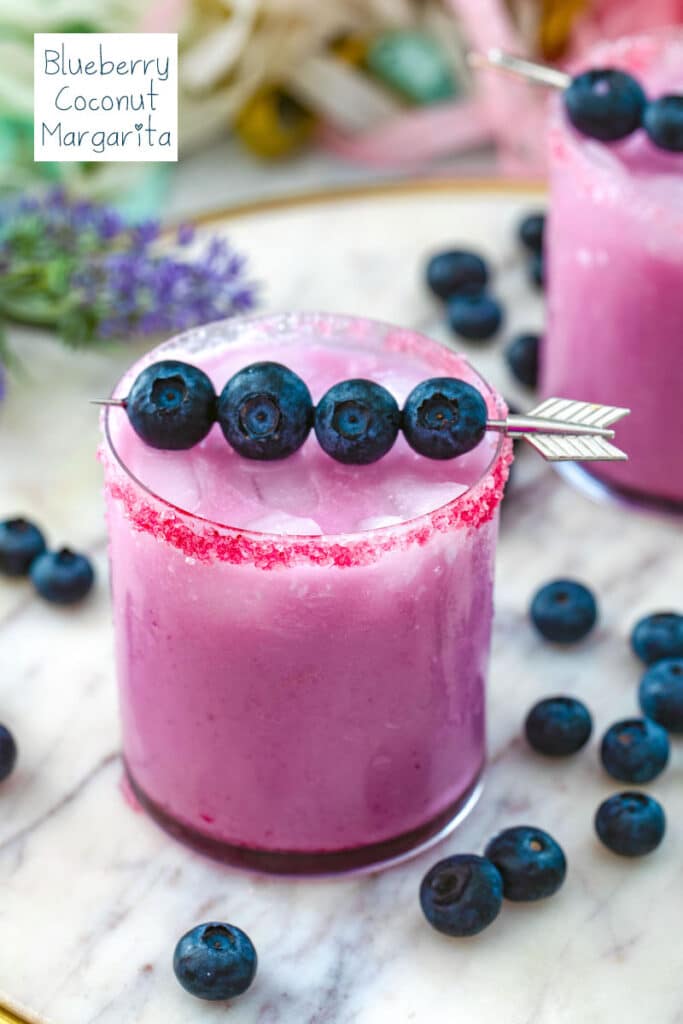 View of a blueberry coconut margarita with fresh blueberry garnish and blueberries in background with recipe title at top