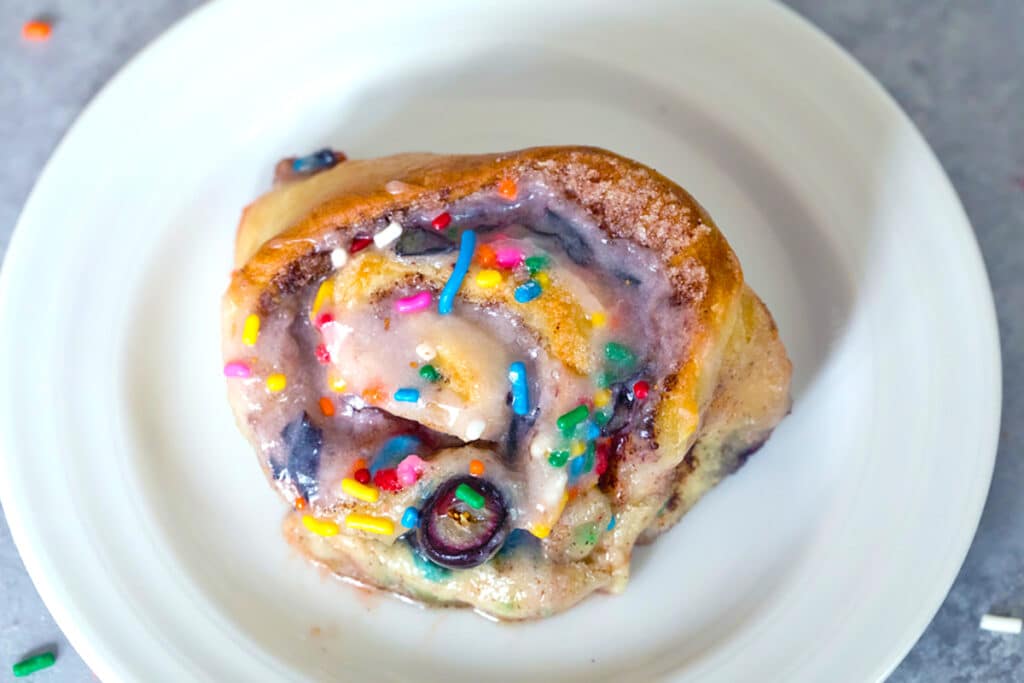 Landscape close-up overhead view of a blueberry funfetti cinnamon roll topped with rainbow sprinkles on a small white plate
