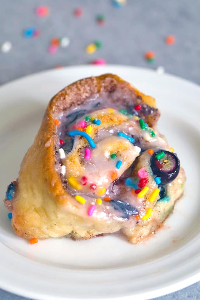 Head-on view of a blueberry funfetti cinnamon roll with rainbow sprinkles and blueberry icing on a small white plate with rainbow sprinkles in the background and recipe title at top