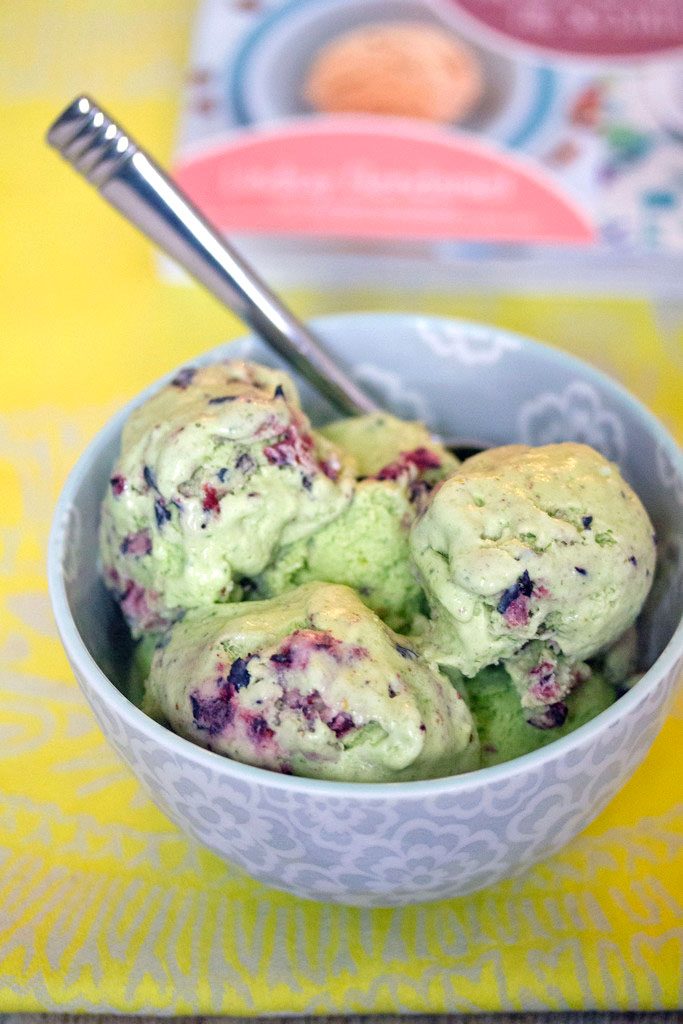 Head-on view of blueberry kale ice cream in a grey bowl with a spoon with cookbook in the background