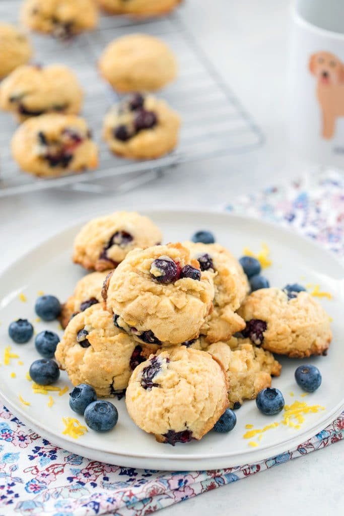 A grey plate on a floral towel with a stack of Blueberry, Lemon, and White Chocolate Chunk Cookies surrounded by blueberries and lemon zest with rack with more cookies and mug in the background