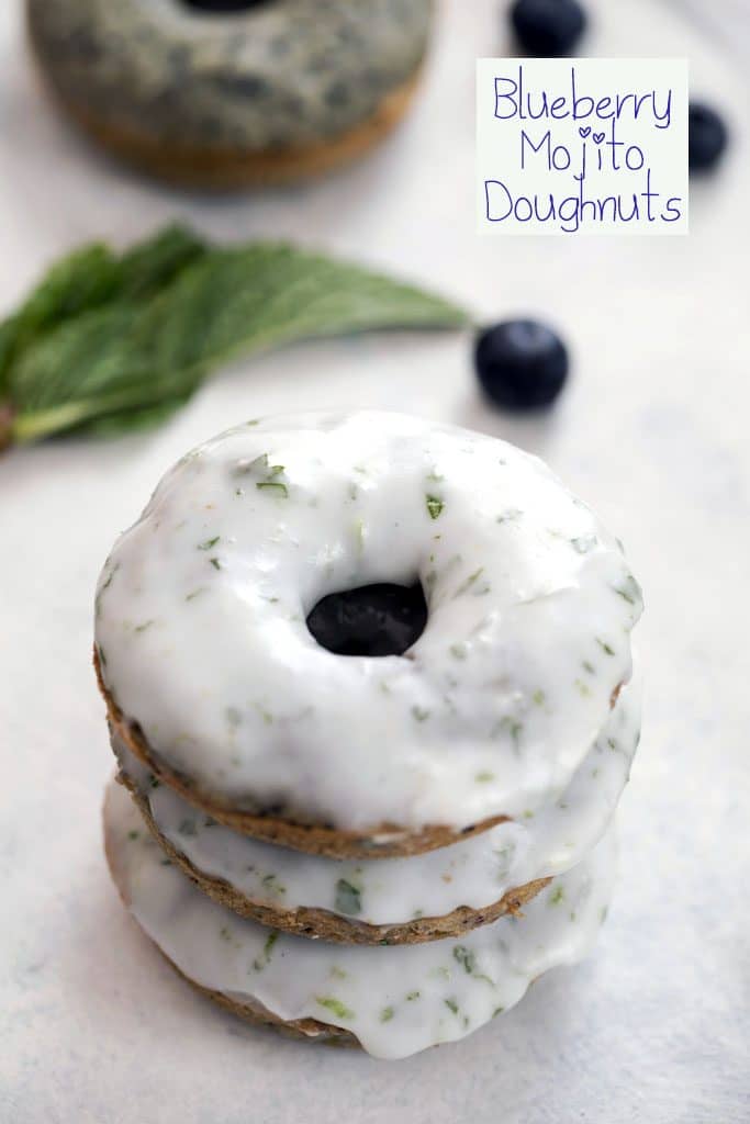 Close-up of a stack of three blueberry mojito doughnuts covered in icing with blueberries and mint in the background and "Blueberry Mojito Doughnut" text at the top