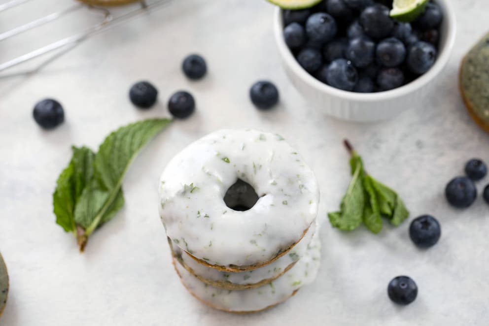 A landscape photo featuring a stack of blueberry mojito doughnuts with a bowl of blueberries in the background, along with blueberries and mint leaves scattered around