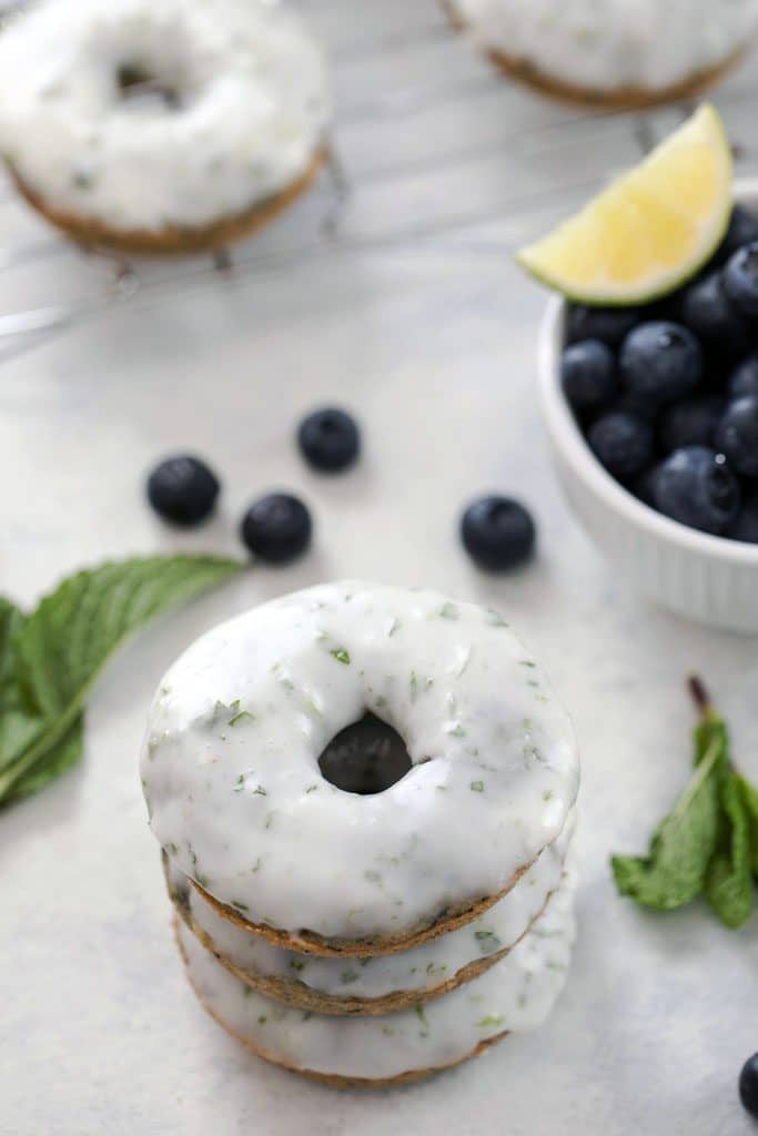 Overhead view of stack of three blueberry mojito doughnuts covered in mint lime icing with a baking rack with more doughnuts in the background, a bowl of blueberries and a lime, and mint