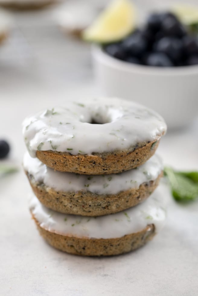 A close-up head-on view of a stack of three blueberry mojito doughnuts covered in mint lime icing with a bowl of blueberries and a lime wedge in the background