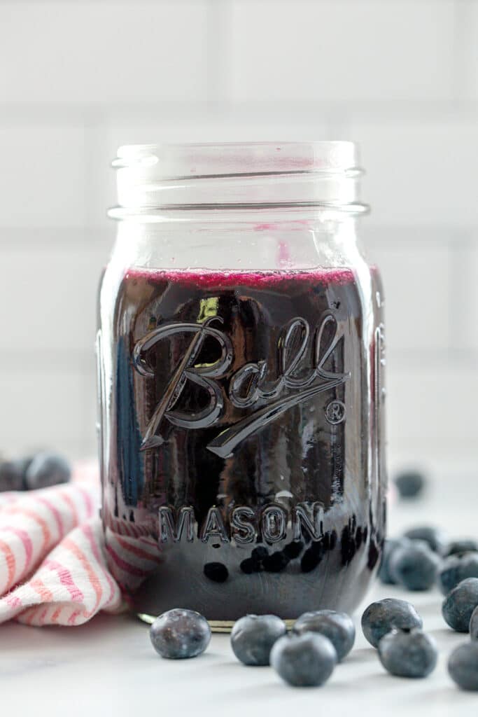 Head-on view of a jar of blueberry simple syrup with blueberries all around and recipe title at top