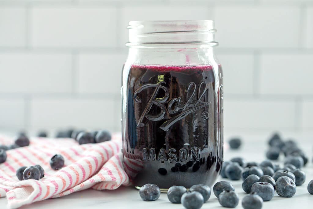 Landscape head-on view of a mason jar of blueberry simple syrup with blueberries all around