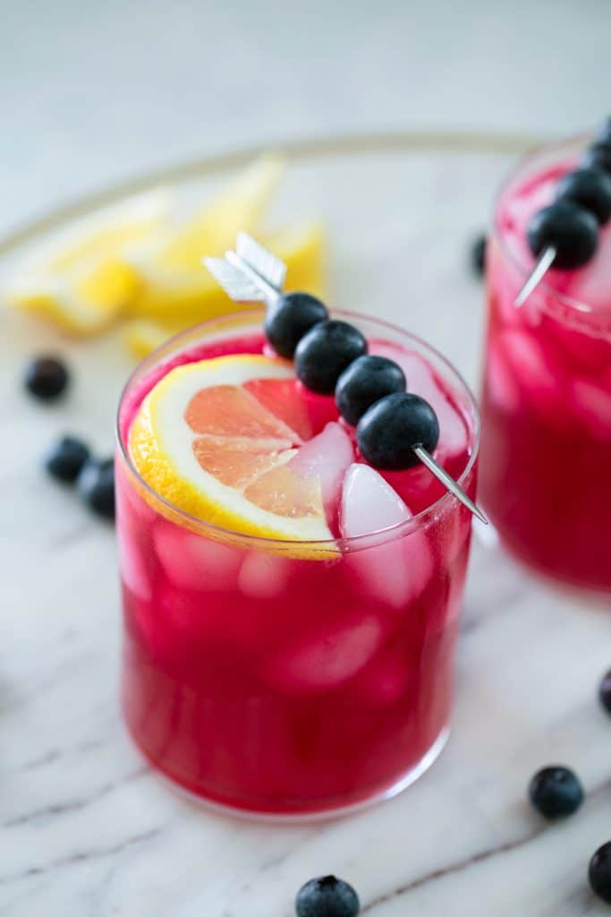 Glass of magenta colored blueberry vodka lemonade in a glass with eyes topped with a skewer of blueberries and a lemon wedge on a marble platter with blueberries and lemons in the background