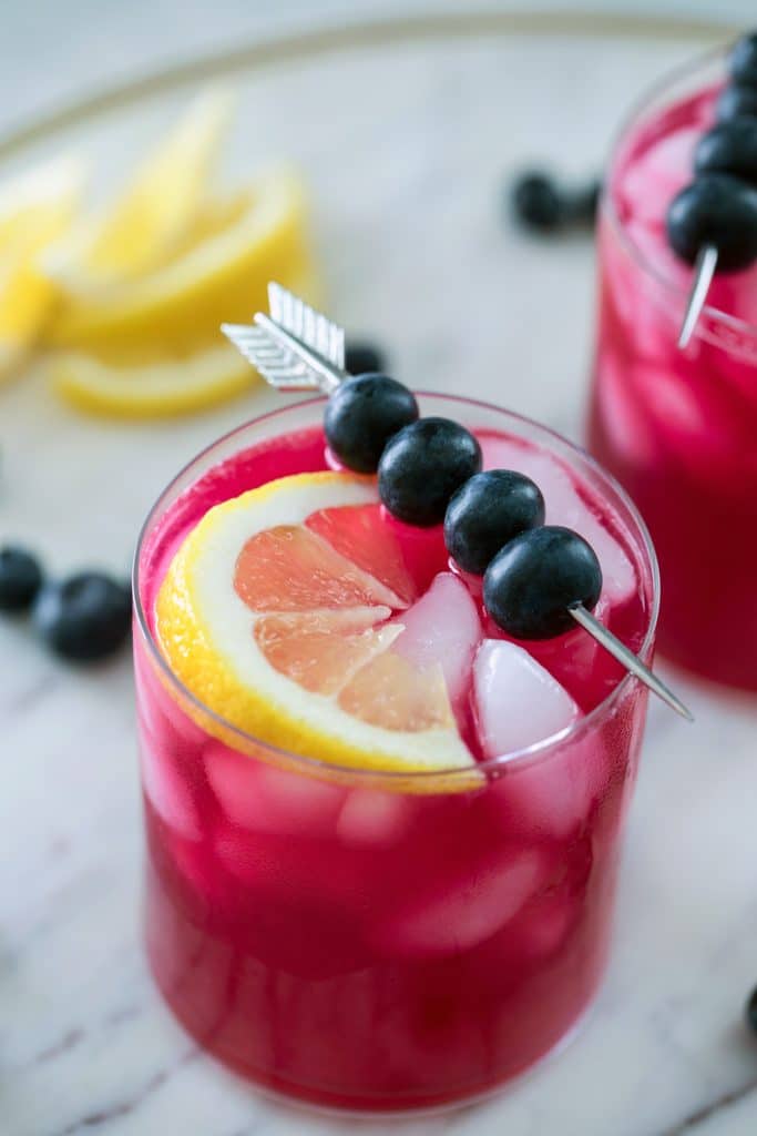 Close-up of a magenta blueberry vodka lemonade cocktail topped with skewer of blueberries and lemon wedge with blueberries and lemons in background on a marble platter