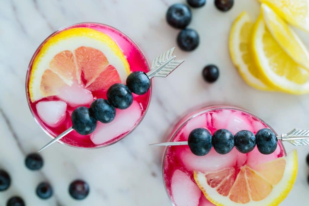 Landscape bird's eye view of two blueberry vodka cocktails topped with arrow skewers of blueberries and lemon wedges on a marble platter with blueberries and lemon wedges in the background