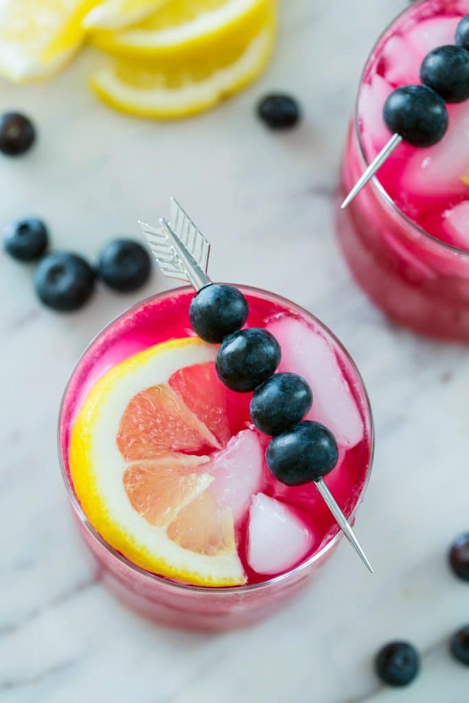 Bird's eye view of magenta blueberry vodka lemonade on a marble platter and topped with arrow skewer of blueberries with blueberries and lemon wedges in the backround